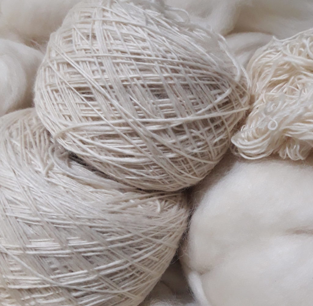 Comecei a Tecer: Como a Tecelagem Mudou Minha Vida — e Pode Mudar a SuaPor que seu fio se desfaz? 8 Benefícios da Arte de Fiar Foto de dois rolos de fios simples (single) torcidos manualmente, esperando para serem equilibrados, juntamente com um chumaço de fibra de lã de carneiro. Esta é a foto ilustrativa do curso de Fiação de lã de carneiro.