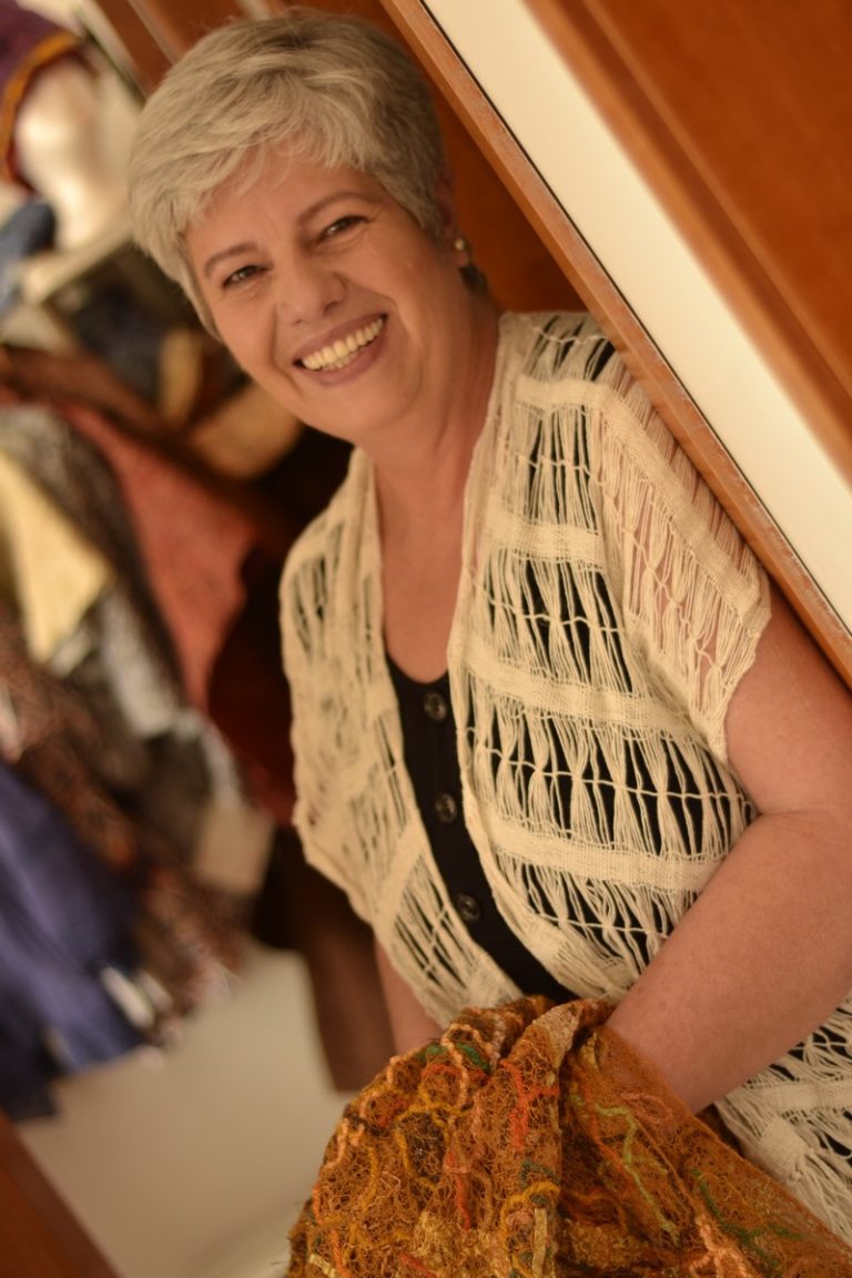 Cursos Isatramas: Foto de Isaura Mattos em pé, com ombro direito encostado no batente de uma porta, sorrindo para você. Isaura Mattos: A Alma Criativa por Trás do Atelier Isatramas. Wrap Isatramas