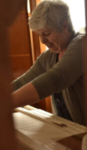 Tecelagem e Fiação: Benefícios para Mente e CérebroQuem ensina transforma. Quem vive o que ensina transforma ainda mais.. 3 Vantagens Neurológicas da Tecelagem Manual. Foto de Isaura Mattos sorrindo e fazendo renda em um tear de pedal. Imagem ilustrativa da Home page do site Atelier Isatramas